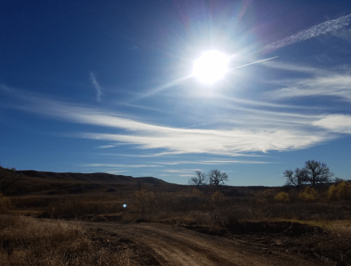 A bright sun shines over a vast landscape with rolling hills and wispy clouds against a clear blue sky.