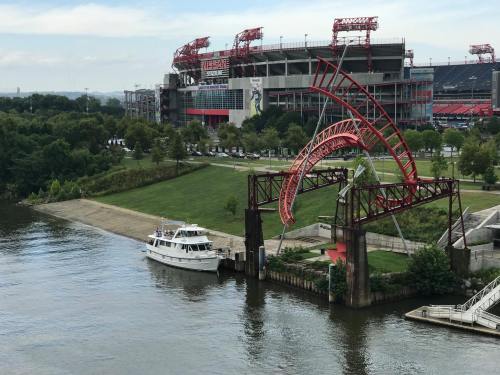 A boat docked by a river with a stadium in the background, featuring red arches and green landscaping.