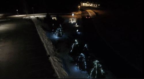 A nighttime scene featuring illuminated trees along a snowy path, with a distant house and streetlights visible.