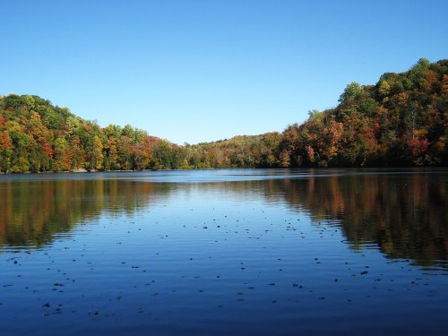 A serene lake surrounded by colorful autumn trees under a clear blue sky, reflecting the vibrant foliage.