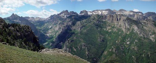 A panoramic view of rugged mountains with green valleys and patches of snow under a blue sky with scattered clouds.