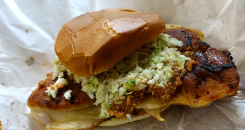 A sandwich with a grilled chicken patty, topped with coleslaw, served in a soft bun on a paper wrapper.