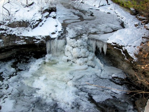 A frozen waterfall cascading into a snowy, icy stream surrounded by rocky terrain and bare trees.