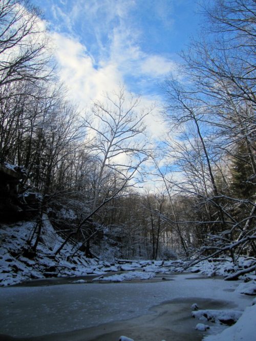 A serene winter landscape featuring a frozen river, snow-covered trees, and a bright blue sky with scattered clouds.