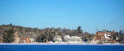 Snow-covered buildings line a lakeshore under a clear blue sky, surrounded by trees in winter.