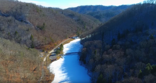 Aerial view of a snowy river winding through wooded hills, with a small cabin near the water's edge.