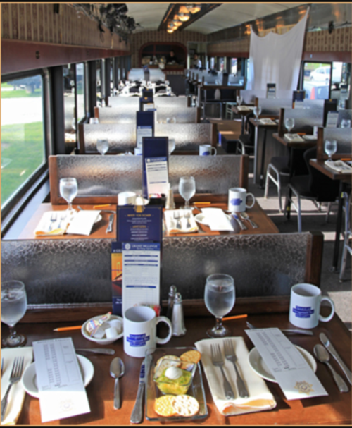 Interior of a dining car with neatly set tables, water glasses, menus, and decorative tableware.