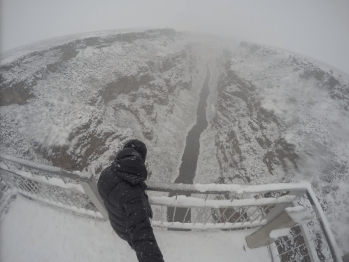 A person stands on a snowy overlook, gazing down at a deep canyon shrouded in fog and falling snow.