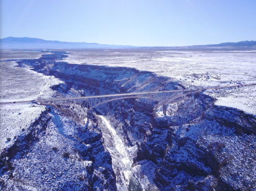 Aerial view of a snowy canyon with a bridge spanning across a deep gorge under a clear blue sky.