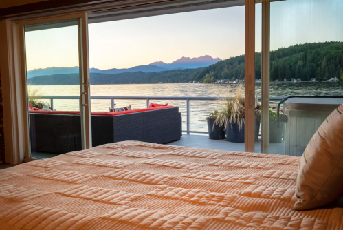 A cozy bedroom with a view of a lake and mountains through sliding glass doors at sunset.