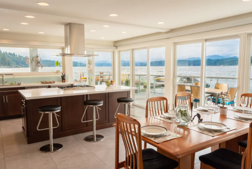 Modern kitchen with a dining area, large windows overlooking a lake, and stylish bar stools. Bright and inviting atmosphere.