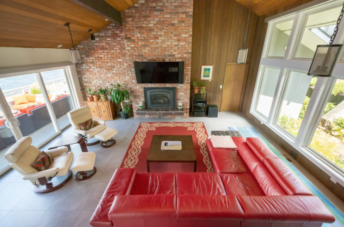 A spacious living room with a red sectional sofa, brick wall, large windows, and a cozy fireplace.