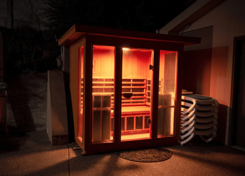 A brightly lit outdoor sauna at night, with wooden benches and glass doors, surrounded by a darkened environment.
