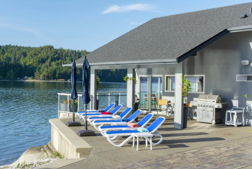 A lakeside patio with lounge chairs, umbrellas, and a grill, surrounded by trees and water under a clear blue sky.
