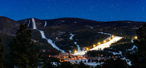 A snowy mountain landscape at night, with ski slopes illuminated and a small village nestled in the valley below.