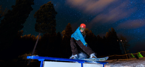 A snowboarder performs a trick on a rail under a starry night sky, surrounded by tall trees.
