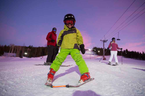 A child in a green ski suit poses on a snowy slope, with skiers and a ski lift in the background at sunset.