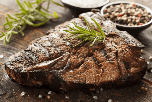A grilled steak garnished with fresh rosemary, surrounded by bowls of salt and pepper on a wooden surface.