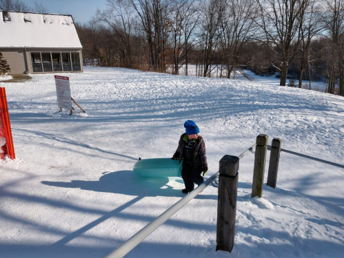 A child in winter gear pulls a blue sled up a snowy hill, with trees and a building in the background.