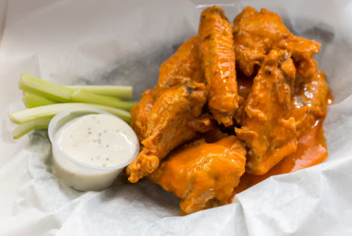 A serving of spicy chicken wings with celery sticks and a side of ranch dressing on a white paper-lined tray.