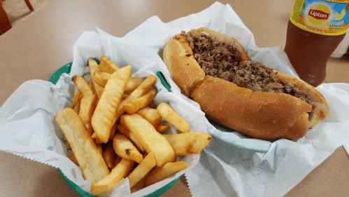 A basket of golden fries next to a large sandwich filled with shredded meat, with a drink in the background.