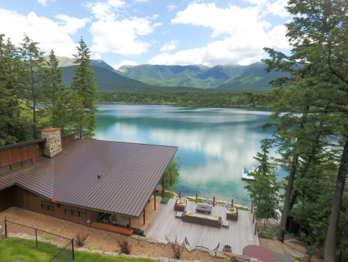 Scenic view of a lakeside house with a deck, surrounded by trees and mountains under a partly cloudy sky.