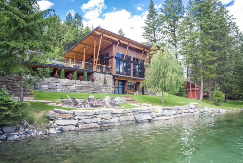 A modern lakeside cabin surrounded by trees, featuring a stone patio and seating area by the water.