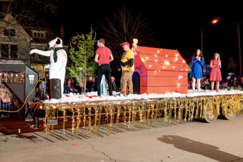 A festive parade float with characters, including a doghouse, decorated with lights and surrounded by people in colorful outfits.