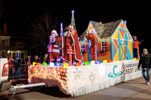 A festive parade float with Santa and performers, decorated with colorful lights and holiday decorations.
