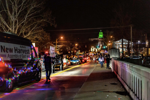 A festive night scene with a parade on a street, decorated vehicles, and people walking, illuminated by colorful lights.