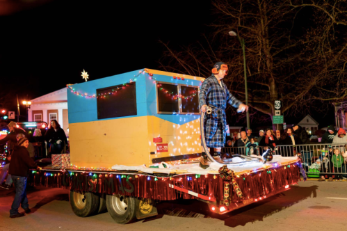 A festive parade float with colorful lights, featuring a person in a robe and hat, surrounded by cheering spectators.