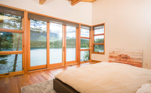 Cozy bedroom with large windows overlooking a serene lake and mountains, featuring wooden accents and a soft bed.