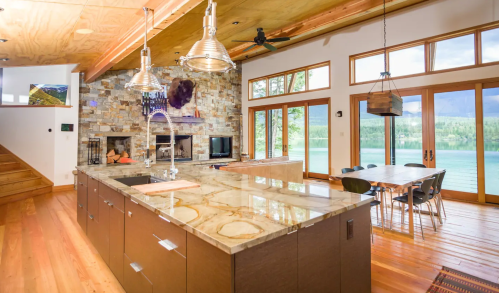 Modern kitchen with a large island, stone fireplace, and dining area, overlooking a scenic lake through large windows.