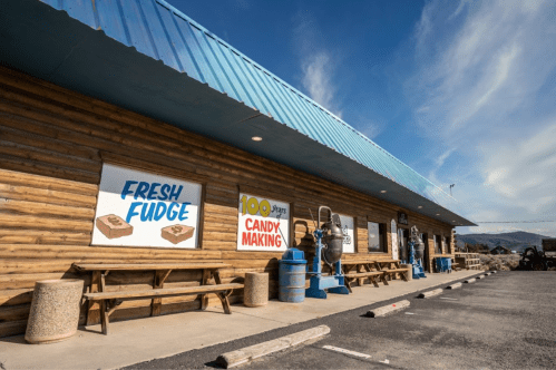 A rustic building with a blue roof, featuring signs for fresh fudge and candy making, surrounded by outdoor seating.