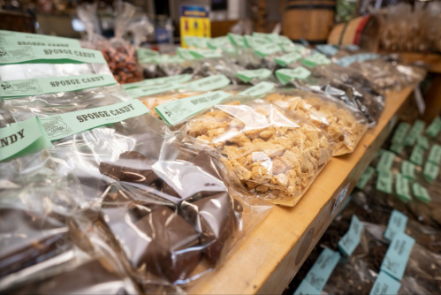 A variety of packaged candies displayed on a wooden table, labeled with green tags, in a market setting.