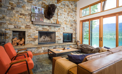 Cozy living room with stone walls, a fireplace, large windows, and a view of the lake surrounded by trees.