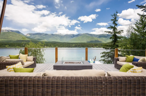 A scenic lakeside view with a cozy seating area, fire pit, and mountains in the background under a blue sky.
