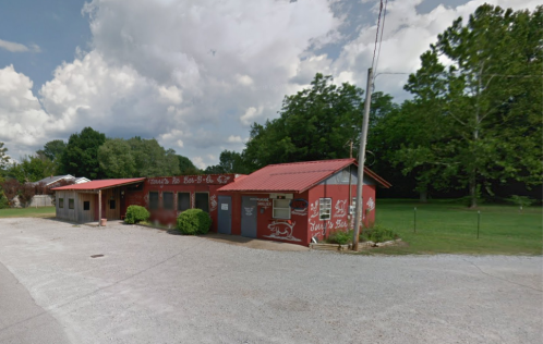 A rustic building with a red roof and painted signage, surrounded by greenery and a gravel road.