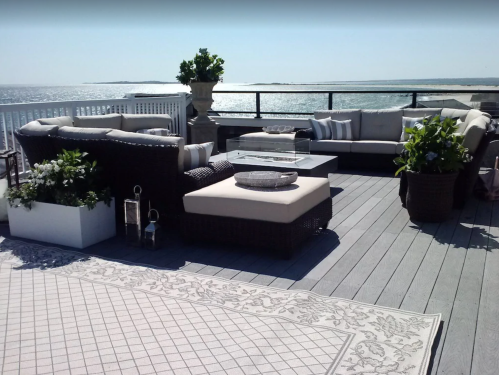 A stylish outdoor seating area with plush furniture, potted plants, and a view of the ocean on a sunny day.