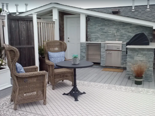 A cozy rooftop patio with two wicker chairs, a small table, and an outdoor kitchen area against a stone wall.