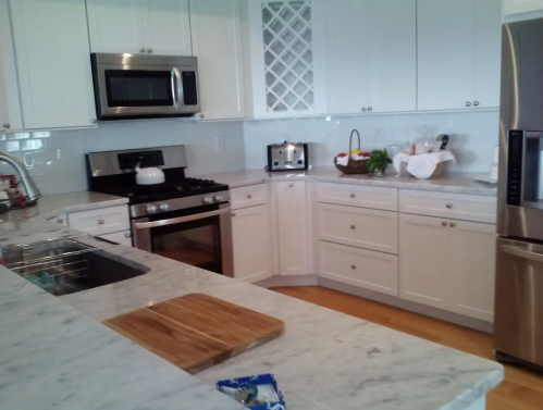 A modern kitchen with white cabinets, stainless steel appliances, and a marble countertop, featuring a cutting board.