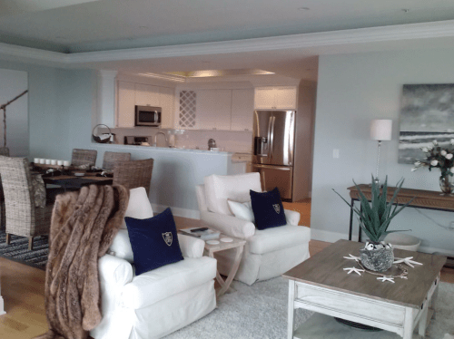 A cozy living area with two white chairs, a wooden table, and a kitchen in the background, featuring light decor.
