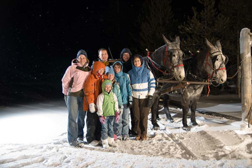 A group of eight people in winter clothing stands beside two horses in a snowy landscape at night.