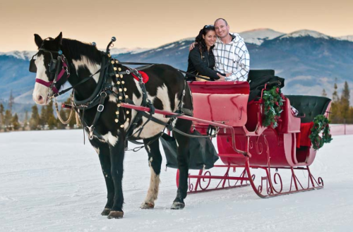 A couple smiles while sitting in a festive red sleigh pulled by a horse, with snowy mountains in the background.