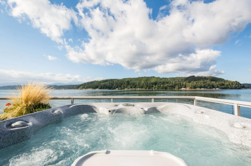 A hot tub overlooking a serene lake surrounded by lush greenery and a blue sky with fluffy clouds.