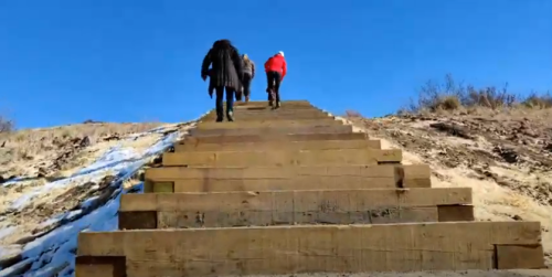 Three people ascend wooden steps leading up a sandy hill under a clear blue sky.