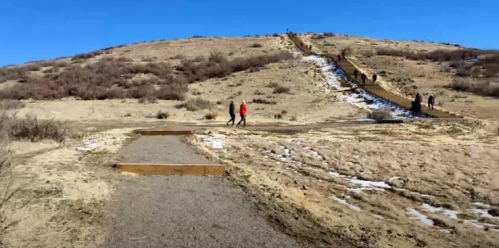 A dirt path leads up a hill with wooden steps, people walking, and sparse vegetation under a clear blue sky.