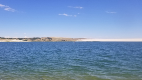A serene view of a blue lake with a sandy shore and hills under a clear blue sky.