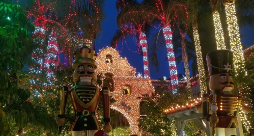 Colorful holiday lights illuminate a garden with nutcracker decorations and palm trees, creating a festive atmosphere.