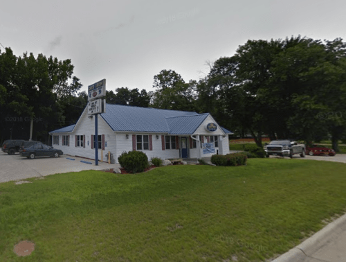 A white building with a blue roof, surrounded by trees and parked cars, featuring a sign in front.